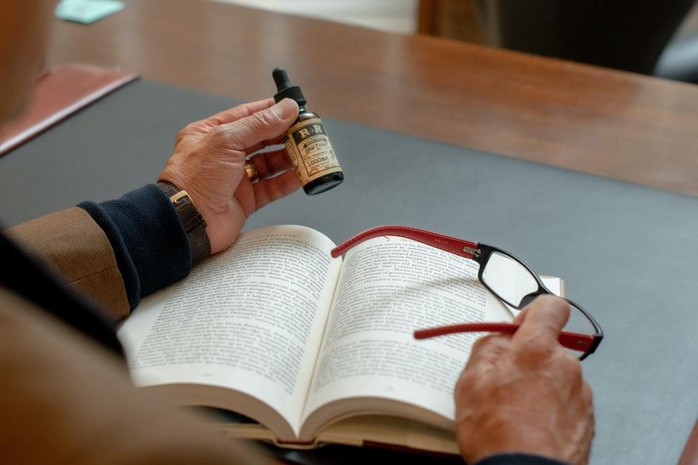 person holding small drop bottle and eyeglasses while reading book